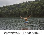 A young guy in blue shorts jumps into a mountain lake and raises his hands up on the background of the forest.                               