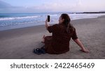 Small photo of Older woman in a smock sitting next to her sandals in front of the sea taking a selfie on the beaches of Cartagena de Indias, Colombia.