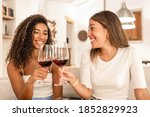 Two young mixed race woman celebrating at home toasting with red wine glasses - Smiling Hispanic curly brunette clinking with her best friend sitting at he sofa in her modern elegant living room