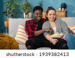 Small photo of Cheerful Caucasian young couple laughing and talking while watching TV together on the couch athome with a bowl of popcorn.