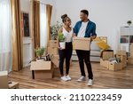 Couple in love moves into new apartment, they carry cardboard boxes with packed belongings into living room, woman sets up decorations, holds pot with plant in hand they decorate first home together