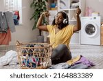 Small photo of Woman of African-American descent sits on floor in middle of laundry room listening to music on wireless headphones while dancing with hands folding clean clothes against backdrop of washing machine.