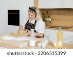 Small photo of Happy elderly woman holding sieve pouring with spoon flour on sieving on wooden table. Spending time in modern kitchen baking cooking homemade cake pizza pasta making surprise for grandchildren.