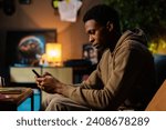 Small photo of Concentrtated African American dark skinned musician, using his phone to record snippets of songs or jot down lyrics while mixing tracks on his laptop.