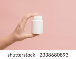 Small photo of White bottle (plastic tube) in hand on a pink background. Packaging for vitamins, pill or capsule, or supplement. Mockup for product branding