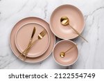 Dishes and utensils for serving and eating meals. Beige round rimmed plates and gold colored cutlery on a white marble table, top view. Modern ceramic crockery, trendy tableware