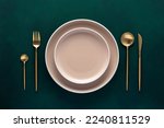 Festive place setting with beige dishes. Empty plate and gold cutlery on dark green background. Dining table in luxury restaurant. Card or menu template, flat design. Tableware, crockery. Top view.