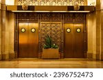 Small photo of Elegant bronze elevator doors with intricate metalwork and art deco patterns, flanked by marble walls and lush greenery in a planter.