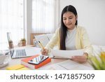 Small photo of Asian female freelancer reading report on laptop and jot down note on table in bedroom at home.Work at home concept.work from home.relax lifestyle