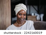 Close up portrait of young woman in turban and white dress sitting on couch at home looking at camera satisfies  by Lise, new home. Beautiful confident African female in head scarf in positive mood.