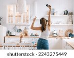 Back view on dancing beautiful Italian girl in headphones, white vest and blue denim jeans shows victory sign bi hand rises hand holds phone. Playful young woman enjoys weekend at home, kitchen.