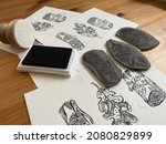 Small photo of A multiple sheets of paper with black and white prints on them, beneath the hand-carved stamps used for the printing, an inkpad and a barren with felt surface.