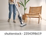 Side view of crop faceless housewife in casual outfit and slippers cleaning laminate floor with modern upright vacuum cleaner in minimalist light apartment