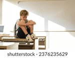 Small photo of Emotionless schoolboy with backpack sitting on desk in classroom at school and embracing knees