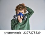 Thoughtful little boy with blond hair in casual clothes putting on protective respirator and looking away against gray background