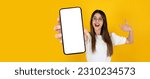 Small photo of Pointing finger mobile phone, portrait of young caucasian excited girl pointing finger mobile phone. Blank empty white big screen smartphone mockup. Isolated yellow studio background, copy space.