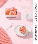 Small photo of A closeup shot of three strawberry snowballs in a porcelain bowl and single one served in a white plate with a saucier and fork on a pink table