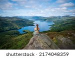 A scenic shot of a woman from her back sitting on a rock on the mountain looking at the calm lake
