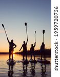 Small photo of Four friends on stand up paddle board (SUP) on a flat quiet winter river at sunset raising his paddles up