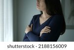 Small photo of Worried woman standing by window touching her neck and chest seeking comfort. Thoughtful preoccupied person