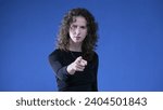 Small photo of Woman accusing viewer by pointing finger at camera while standing on blue background. Upset doubtful person gesturing at offender
