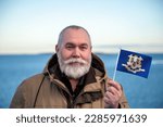 Small photo of Man holding Connecticut state flag. Portrait of older man with Connecticut flag. Visit Connecticut. Older man 50 55 60 years old with gray beard outdoors travelling.