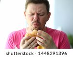Small photo of Portrait of unsatisfied male eating tasteless hamburger with obvious disgust. Bearded person holding hideous and sickening burger in hands. Blurred background