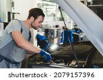 Small photo of Caucasian young male car technician mechanic in special blue robe uniform holding pipe monkey wrench and flash light repairing fixing car at vehicle service inspection. MOT