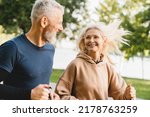 Mature middle aged senior couple running together in the park stadium looking at each other while jogging slimming exercises. Training workout