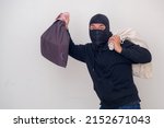 A male thief in a black mask and hood carries two sacks of stolen goods and some money.