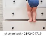 Small photo of Toddler baby climbed up on the open chest of drawers. Child boy stood on a tall drawer of a white cabinet. Kid age one year