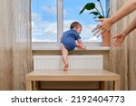 Small photo of Toddler baby climbs to the window and the hands of a frightened mother. Child crawls to the window holding on to the furniture in the home living room. Kid age one year