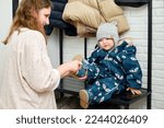 The mother puts a blue shoes on the foot toddler baby sitting in the home hallway. Woman mom dressing warm boots clothes on child for winter walk in cold weather. Kid aged one year and three months