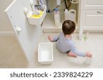 A child is playing with chemical cleaning products under the sink in the kitchen. Baby holds bottles with detergent. Kid aged about two years (one year nine months)