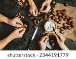 Close up of hands of three people peeling roasted chestnuts on black table background. Lifestyle family meal concept. Dark low key photo. Top view.