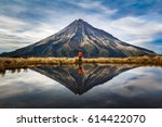 A mountaineer with an orange dress in a symmetric picture looking to the taranaki volcano in the north island of new zealand and with the reflection of tho mountain and the climber in a lake. Zeland