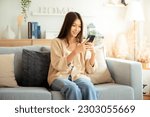 Happy young asian woman relax on comfortable couch at home texting messaging on smartphone, smiling girl use cellphone chatting, browse wireless internet on gadget, shopping online from home