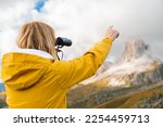 Blonde woman looks at Passo Giau pass through binoculars pointing finger on rocky mountain. Female tourist enjoys hiking in Italian Alps slow motion