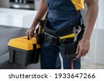 Home repair. Dark-skinned service man standing in the kitchen and holding a box with tools