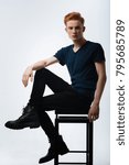 Small photo of Seriousness. Good-looking humourless red-haired young man sitting on the chair and having his hand on his knee and wearing a black t-shirt and trousers and boots