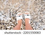 Small photo of Legs of a girl by the sea. Feet in funny slippers on pebbles. Sea wave and slippers. Slippers in the form of kittens.