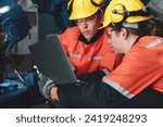 Two Caucasian engineers in hard hats and high-visibility clothing examine industrial equipment using a laptop