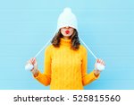 Happy cool girl blowing red lips makes air kiss wearing a knitted hat, yellow sweater over blue background