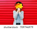 Fashion autumn portrait woman covering her face with yellow maple leaves on a red background