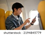 Small photo of side view, A good looking Asian male traveler in jean jacket and headphones sits at the window seat in economy class, using his mobile phone to check his email before takeoff. Airplane journey concept
