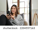 Small photo of Attractive and happy young Asian female using her smartphone to scroll through social media while relaxing on her comfortable sofa in her living room. 