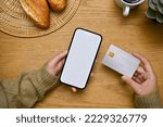 A female hands holding a smartphone white screen mockup and a credit card over wooden tabletop. top view