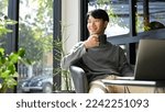 Small photo of Happy and handsome Asian male enjoys having morning coffee at the coffee shop, looking away from camera, daydreaming about his career success.