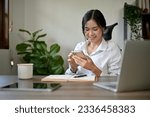 A beautiful Asian businesswoman texting with her friends or checking emails on her smartphone while sitting at her desk in her office.