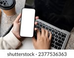 Close-up image of a woman using her smartphone while working on her laptop at her modern desk. A white-screen smartphone to display your graphic ads. People and technology concepts